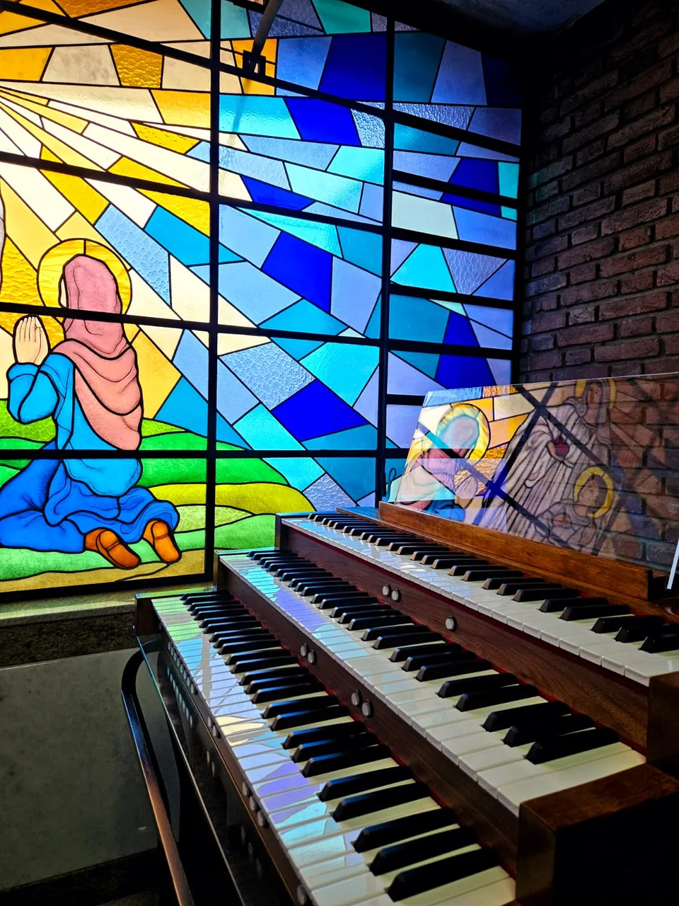 Órgão digital de três teclados em madeira, junto a um vitral colorido.